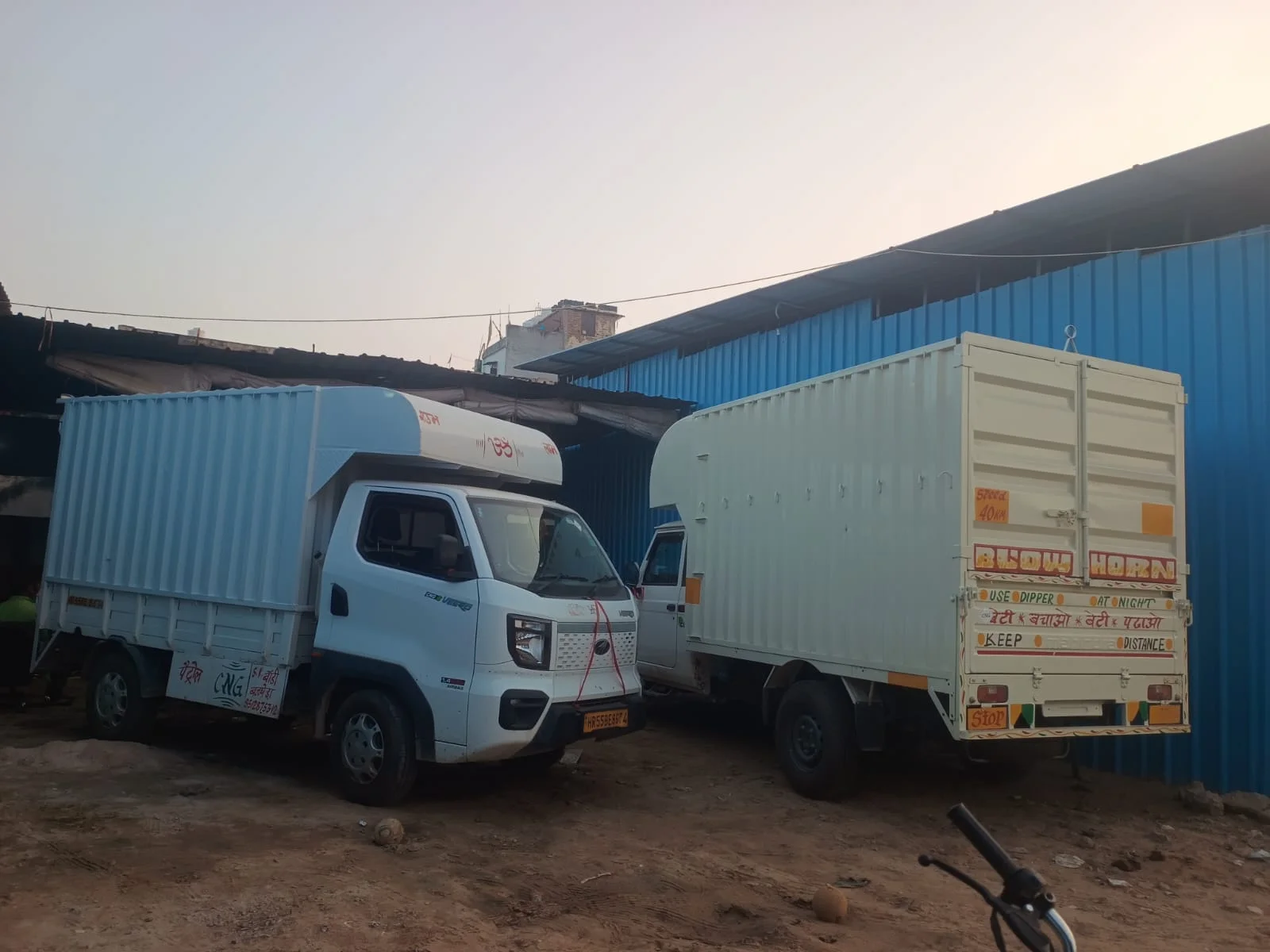 Two small closed-body goods trucks parked beside a transport shed